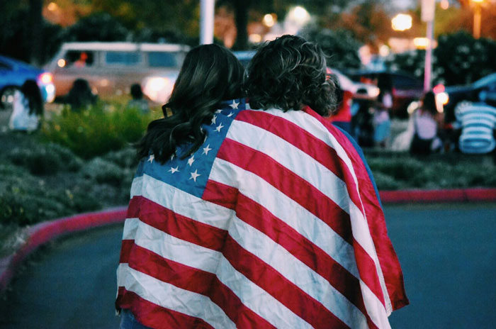Woman wrapped in American flag, experiencing life in a blue state, surrounded by a casual, outdoor gathering.