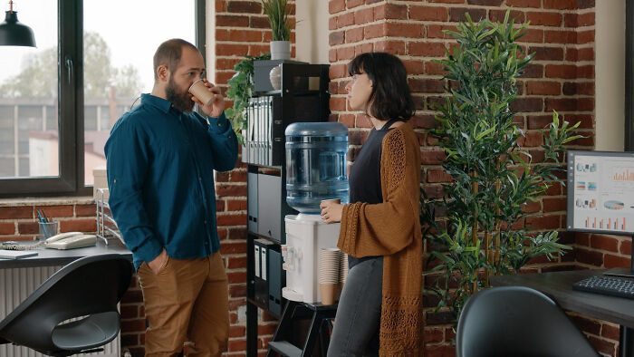 Two colleagues chatting by a water cooler in a modern office, surrounded by plants and equipment.