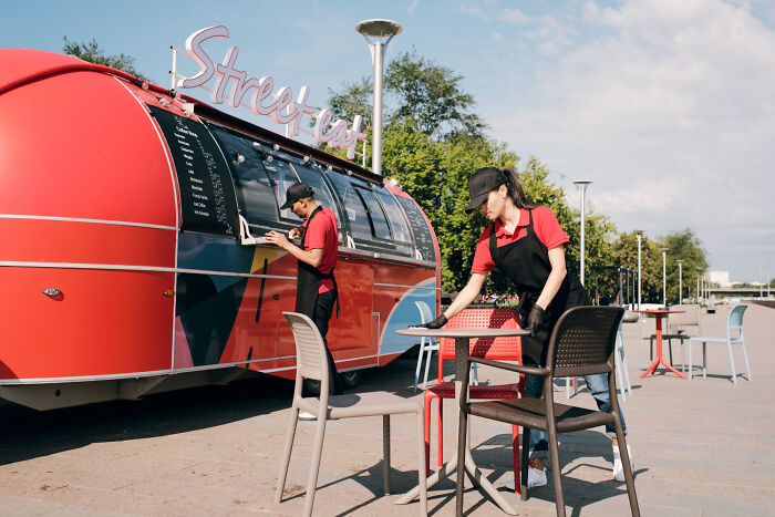 Street food employees in uniforms working at a colorful food truck and setting up tables outdoors.
