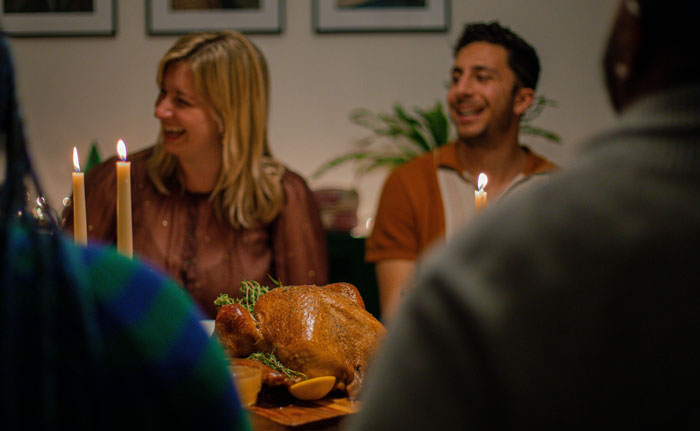 People laughing around a dinner table with a roasted turkey, candles adding warm light to the scene. People laughing around a dinner table with a roasted turkey, candles adding warm light to the scene.