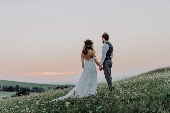 Bride and fianc&eacute; holding hands in a field at sunset, highlighting wedding attire and nature.