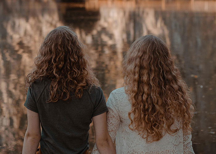 Two women with curly hair sit by a serene lake, highlighting financial choices and family dynamics.
