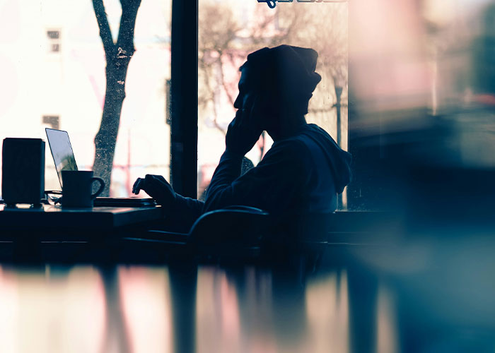 Person in a cafe, silhouette against a window, using a laptop and phone, possibly sharing secrets online.