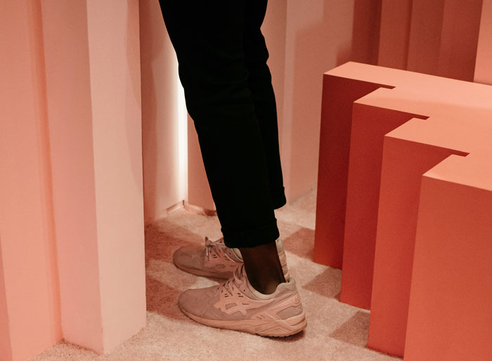 Person wearing white sneakers and black pants, standing among pink geometric pillars, fashion trends critique context.