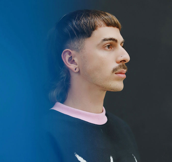 Person with a mustache and mullet hairstyle against a dark background, representing a fashion trend some people dislike.