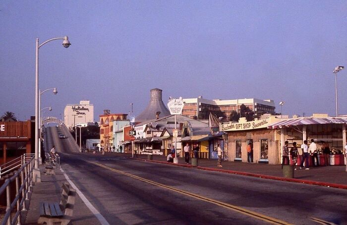 Vintage 1970s West Coast street scene with shops, a bridge, and people strolling on a sunny day.