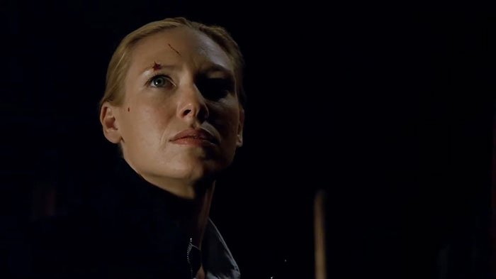A determined woman with a scar on her forehead stands in a dimly lit setting, embodying a strong female protagonist.