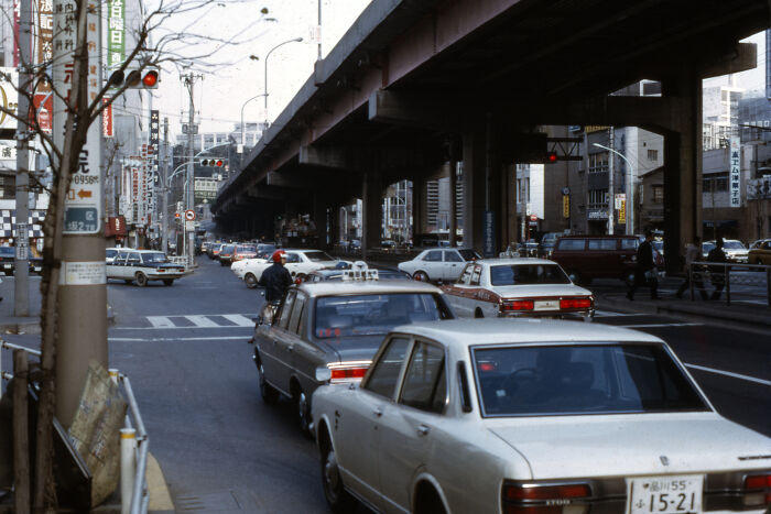 1973 city street scene with vintage cars, elevated highway, and urban landscape in the background.