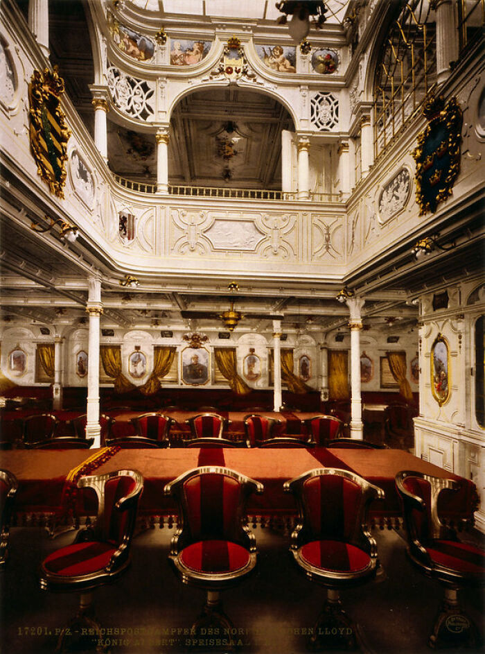 Old color photo of a lavish, ornate ship's dining hall from 100 years ago with red upholstered chairs and decorative walls.