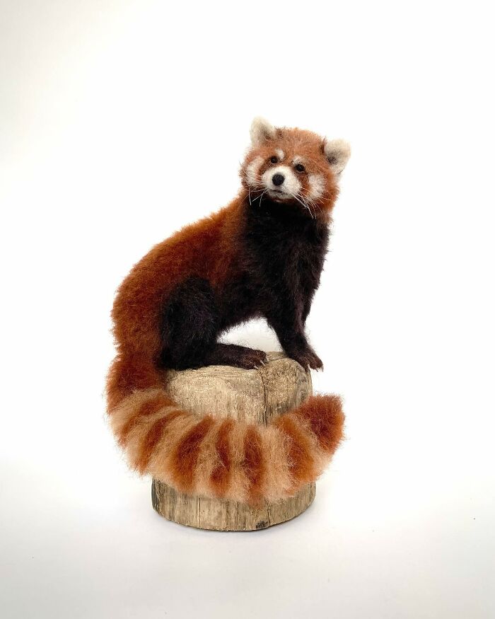 Needle-felted red panda sitting on a wooden stump, showcasing intricate craftsmanship in a charming pose.