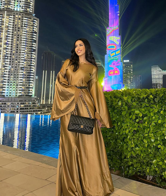 Dubai housewife in gold dress, standing by a pool at night with city lights in the background.
