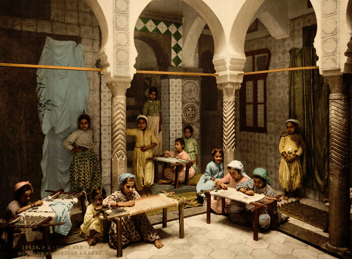 Historic color photo depicting people working in an ornate room 100 years ago, showcasing cultural attire and architecture.