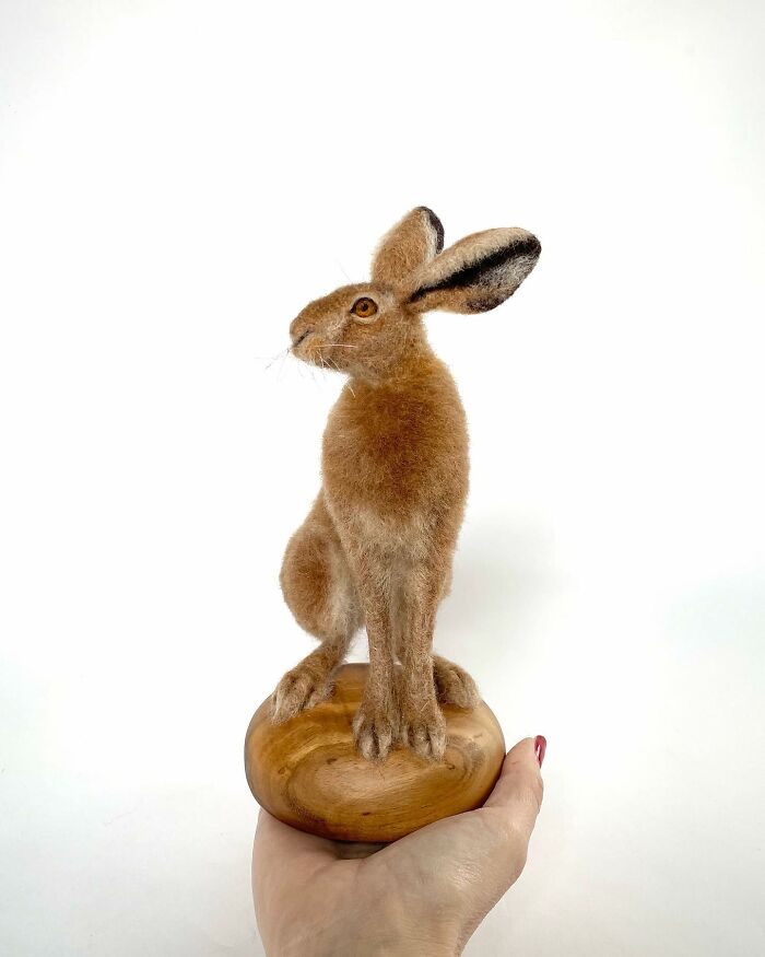 Needle-felted rabbit sculpture on a wooden base, held in a hand, showcasing realistic animal art.
