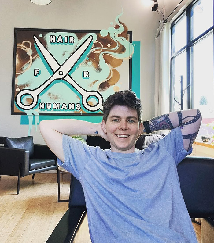 Jules Heron smiling in salon with sign "Hair for Humans," creating an inclusive, safe space for haircuts.