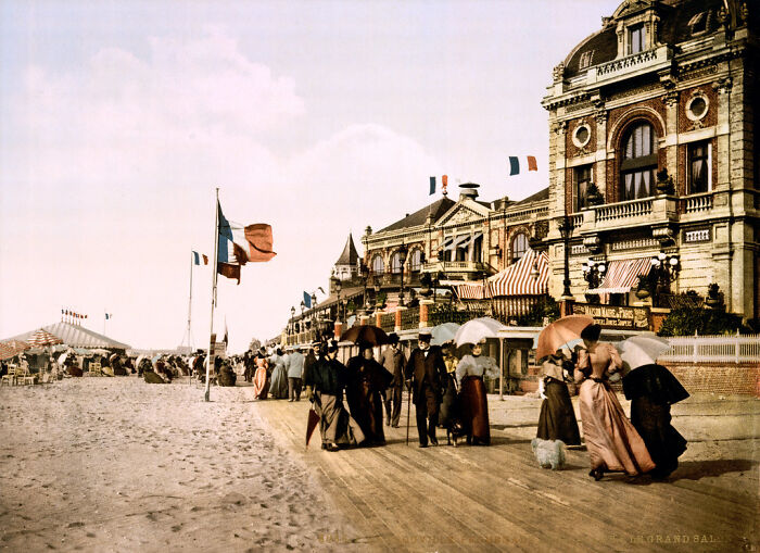 Beach promenade with people in vintage clothing, French flags, and historic architecture, showcasing colors from 100 years ago.