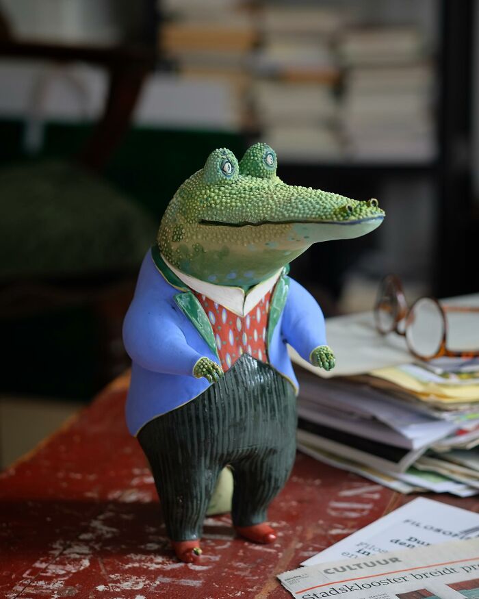 Playful ceramic sculpture of an alligator in a colorful outfit by Nastia Calaca, standing on a table with papers around.