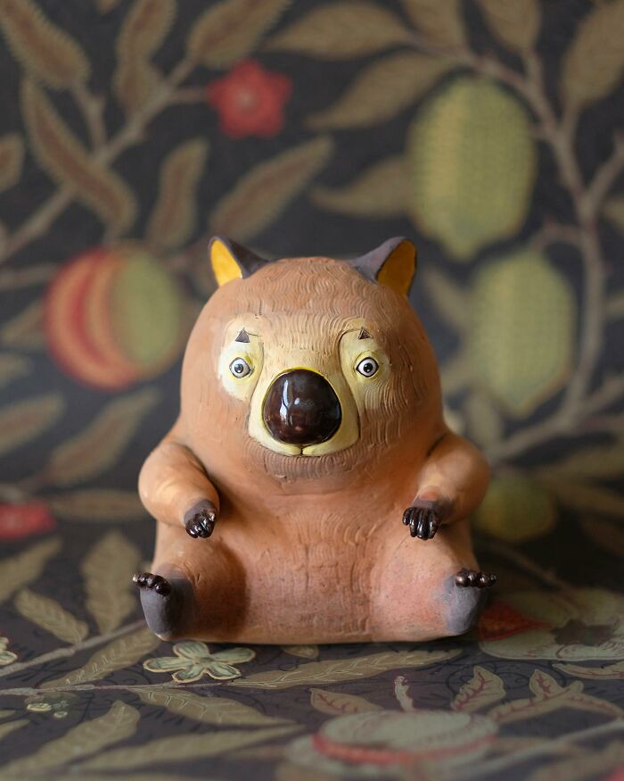 Playful ceramic sculpture of a bear-like figure by Nastia Calaca, set against a floral patterned background.