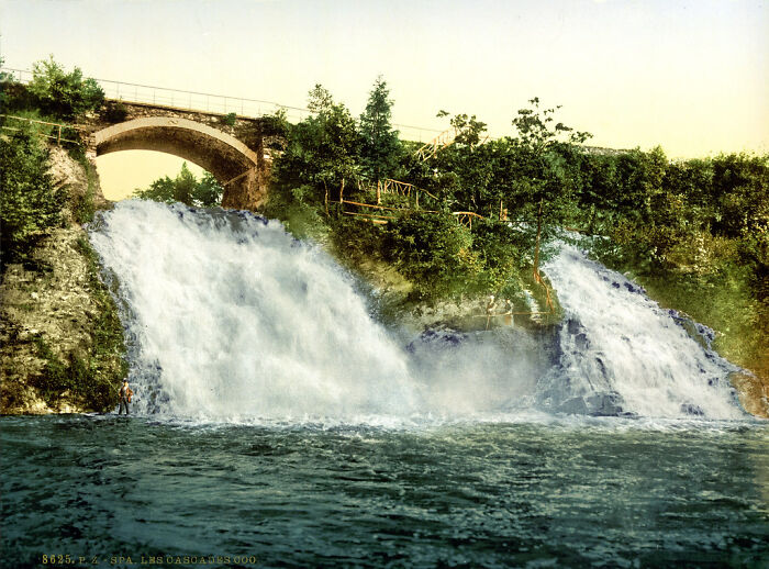 Historic color photo of a waterfall with an arched bridge above, surrounded by lush greenery, from 100 years ago.