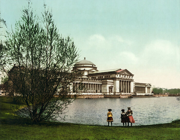 Early color photo of a historical building by a lake, with three children in the foreground, depicting life 100 years ago.