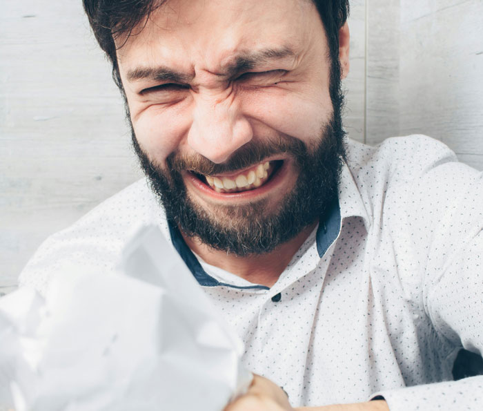 Bearded man in frustration, grimacing with eyes closed, wearing a white shirt, gripping a crumpled paper.