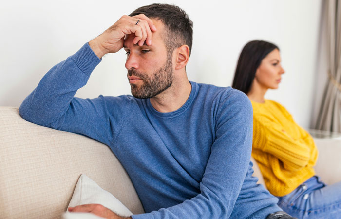 Man in a blue sweater looking upset on a couch, with a woman in a yellow sweater turned away, symbolizing marital conflict. Man in a blue sweater looking upset on a couch, with a woman in a yellow sweater turned away, symbolizing marital conflict.
