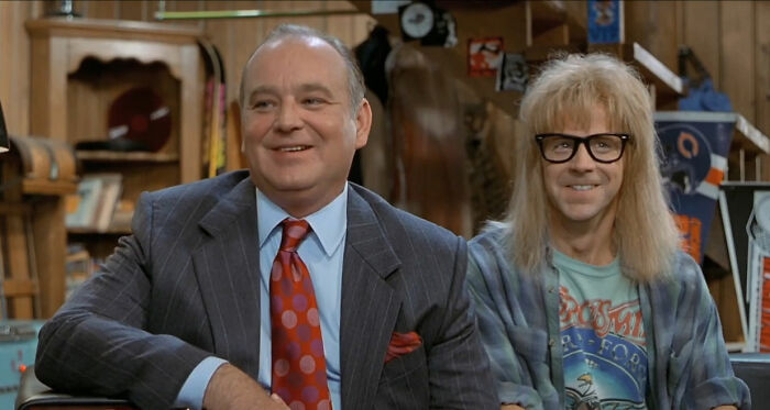 Two characters in a wood-paneled room, one in a suit, the other with long hair and glasses, evoking a movie joke moment.