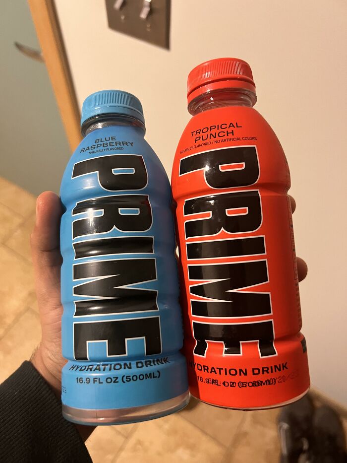 Two PRIME hydration drinks in a person's hand, flavors: Blue Raspberry and Tropical Punch.