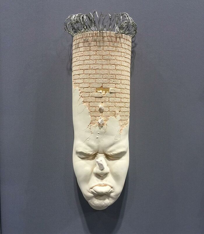 Surrealism and realism merge in a sculpted face with a c*****d brick texture and wire details on top.