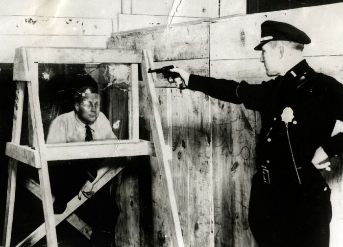 Policeman testing bulletproof glass, a strange invention, by firing a gun at a man behind it.