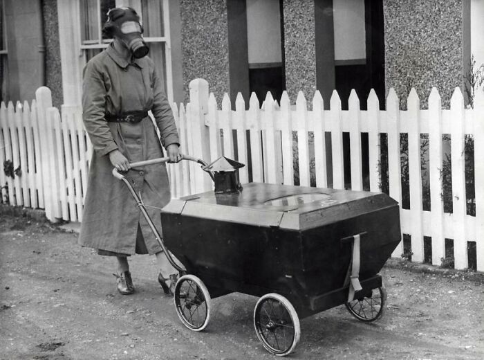 Person in a gas mask pushing a bizarre, box-shaped stroller outside a house.