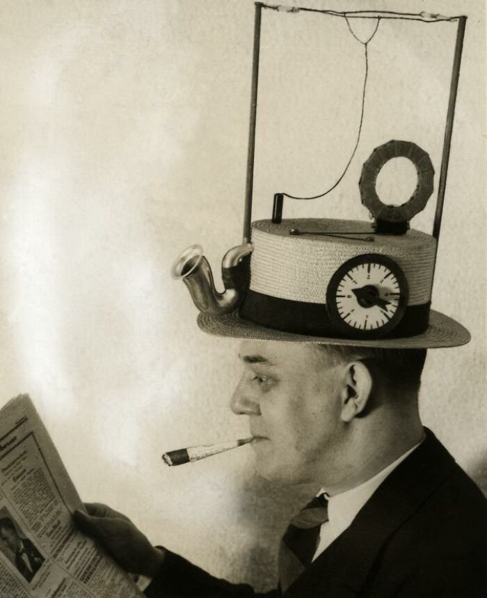 Man wearing a peculiar hat with pipes and a clock, reading a newspaper; example of strange inventions.