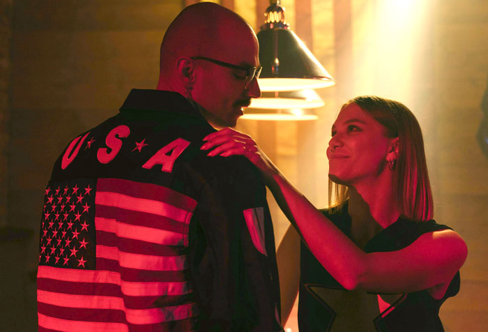 Two people in a dimly lit room, one wearing a USA flag jacket, exemplifying controversial fashion trends.