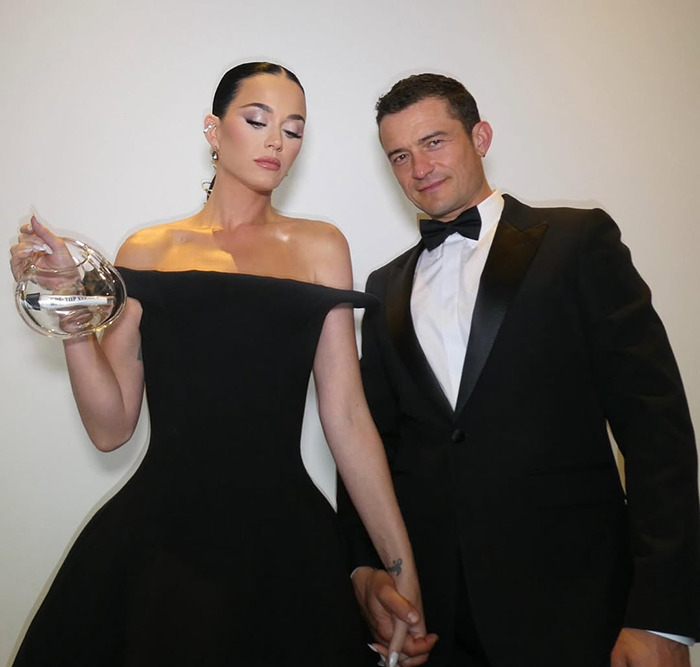 Katy Perry in black dress standing beside partner Orlando Bloom in a tuxedo, holding a clear award. Katy Perry in black dress standing beside partner Orlando Bloom in a tuxedo, holding a clear award.