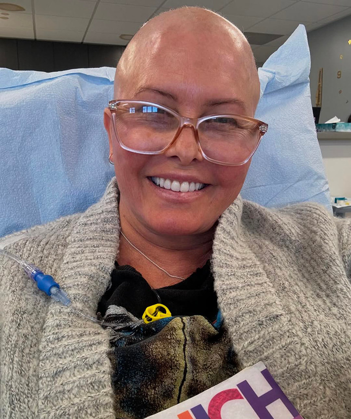 Nicole Eggert in hospital setting smiling, wearing glasses and cozy sweater, highlighting cancer journey.