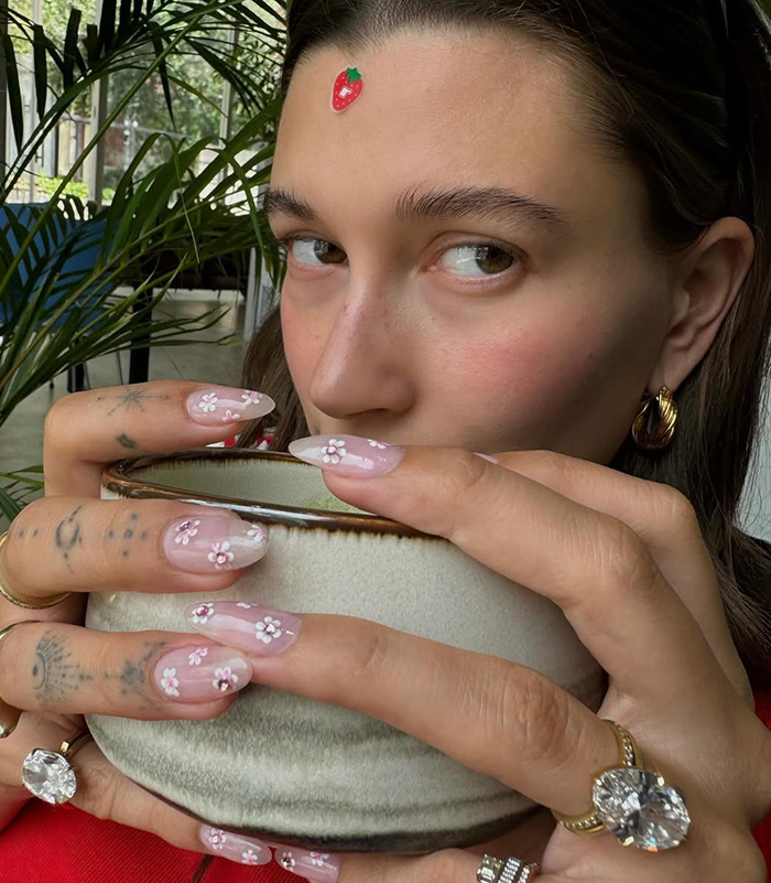 Hailey Bieber holding a cup, showing a diamond engagement ring, with floral nail art and a strawberry sticker on the forehead. Hailey Bieber holding a cup, showing a diamond engagement ring, with floral nail art and a strawberry sticker on the forehead.