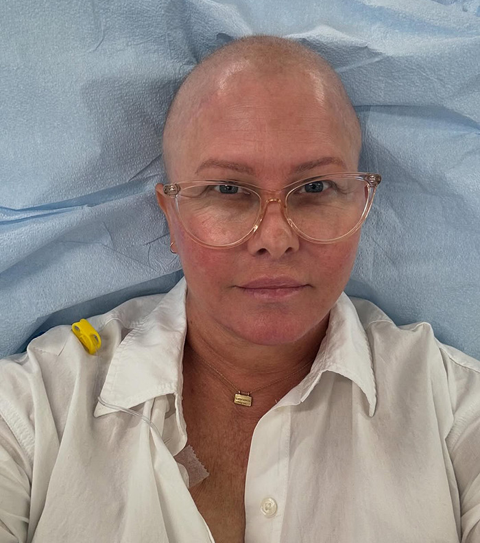 Nicole Eggert wearing glasses and a white shirt, resting on a blue surface, hints of medical setting.