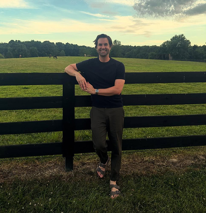 Man smiling and leaning on a black fence in a green field.