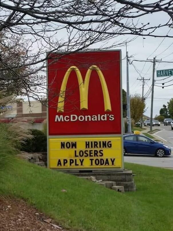Weird and absurd sign at McDonald's reads "Now Hiring Losers, Apply Today" on a busy street corner.
