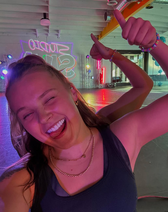 JoJo Siwa smiling and giving thumbs up in a colorful dance studio, related to the celebrity &ldquo;ignore list&rdquo; trend.