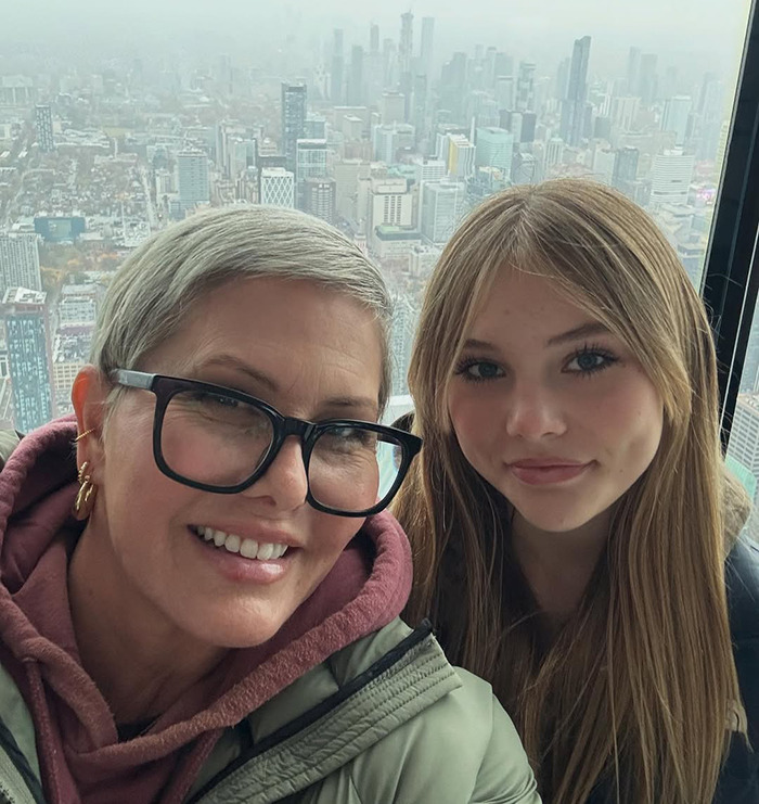 Nicole Eggert with short hair and glasses, posing with her daughter against a city skyline background.