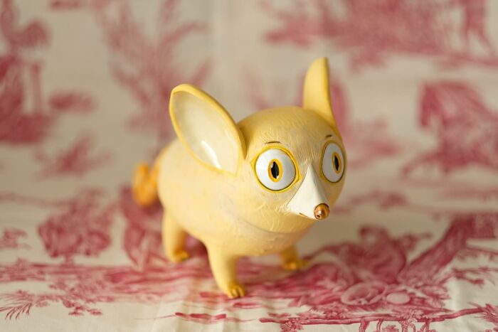 Playful ceramic sculpture by Nastia Calaca featuring a quirky yellow creature with big eyes and ears on a patterned background.