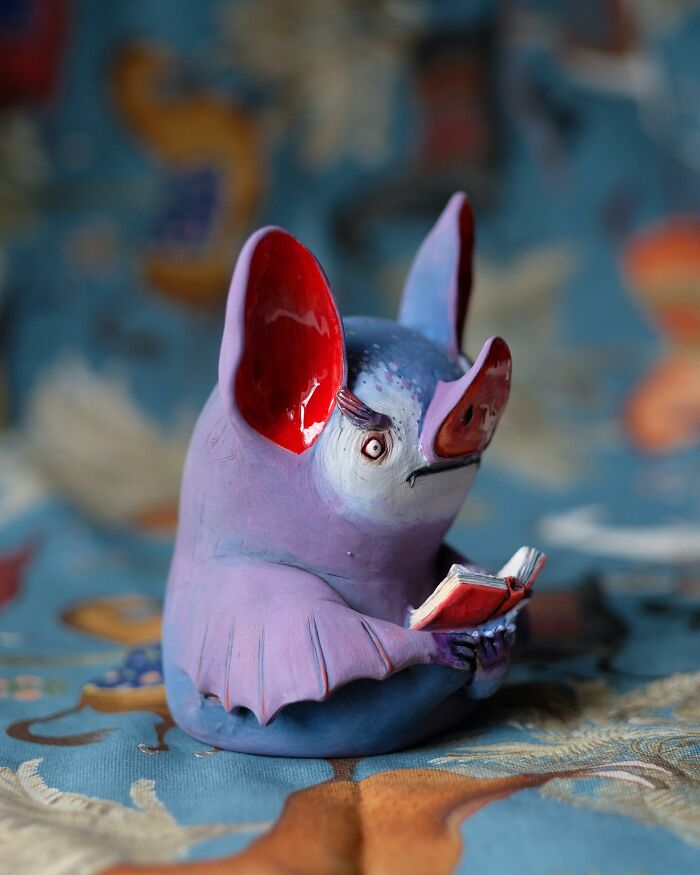 Playful ceramic sculpture of a colorful bat reading a book by Nastia Calaca.