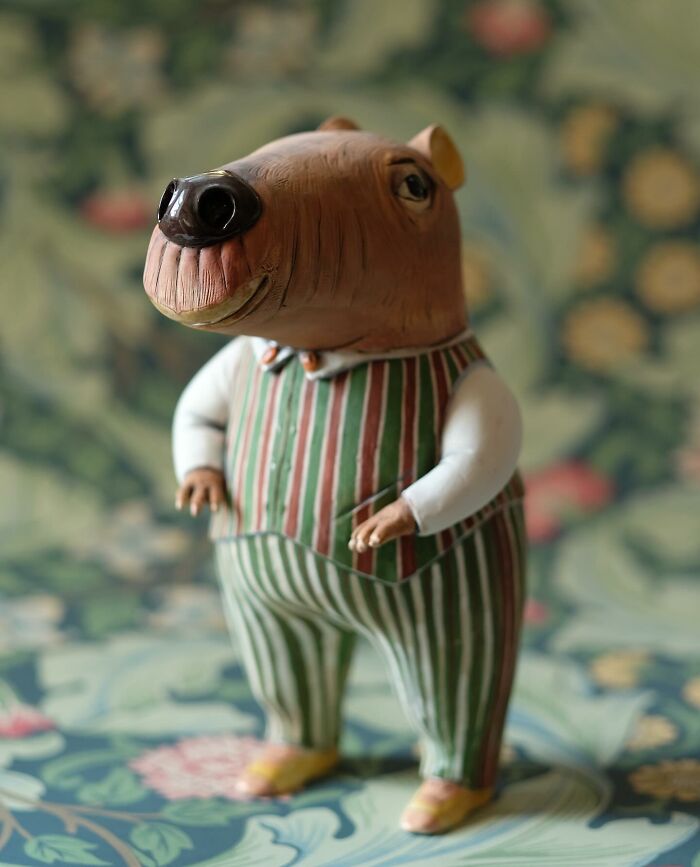 Playful ceramic sculpture of a capybara figure by Nastia Calaca, wearing a striped outfit against a floral background.