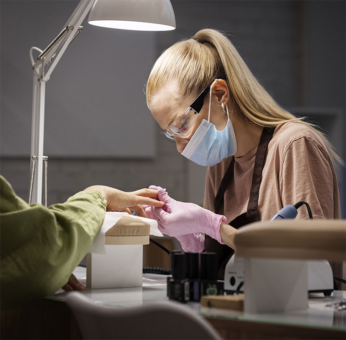 Woman providing nail service, wearing gloves and mask, focused on the task under bright lamp. Woman providing nail service, wearing gloves and mask, focused on the task under bright lamp.
