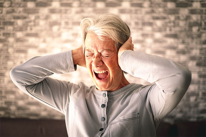 Older woman with grey hair and light shirt, showing stress by covering ears and closing eyes with a pained expression.
