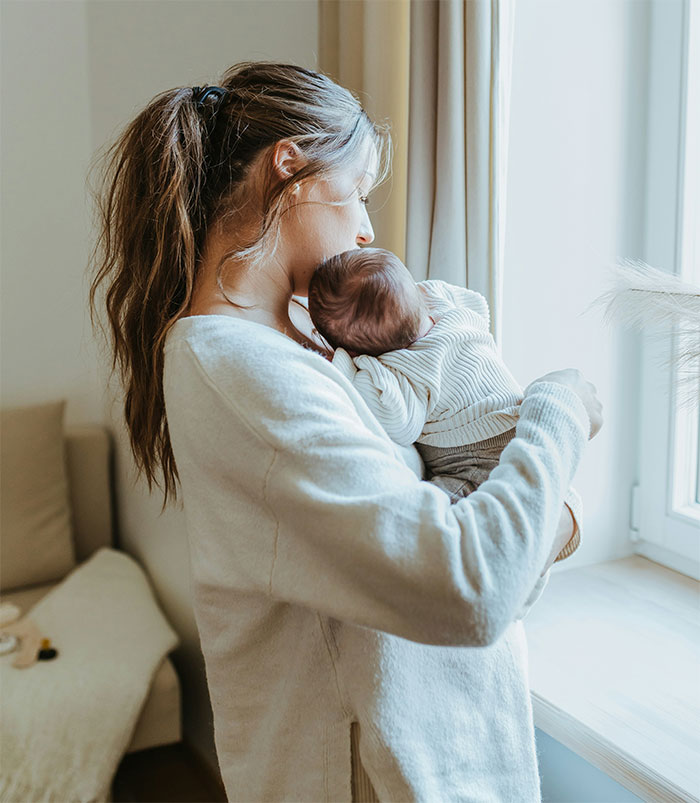 Woman holding baby by a window, wearing a light sweater, showcasing a moment of tenderness and care. Woman holding baby by a window, wearing a light sweater, showcasing a moment of tenderness and care.