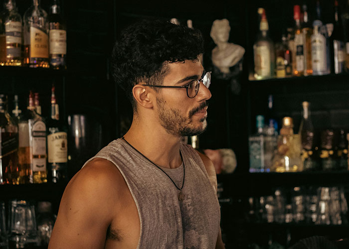 Man at a bar surrounded by bottles, reflecting secrets spilled.