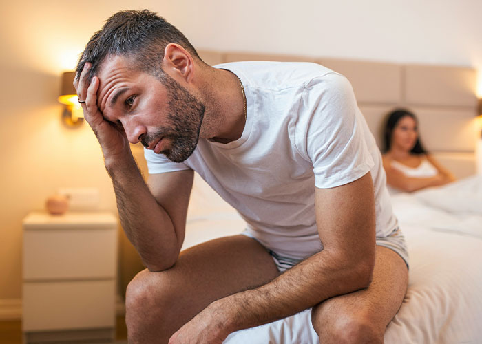 Man looking regretful, sitting on bed, hand on forehead, with a woman in the background.