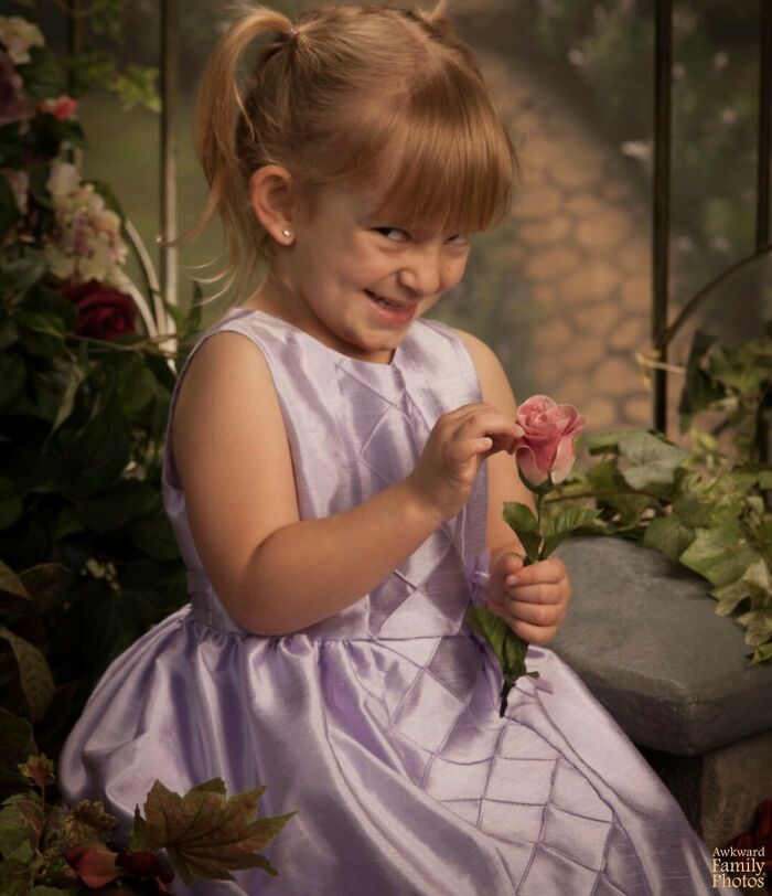 A young girl in a lilac dress smirking while holding a pink rose, showcasing awkward family photo vibes.