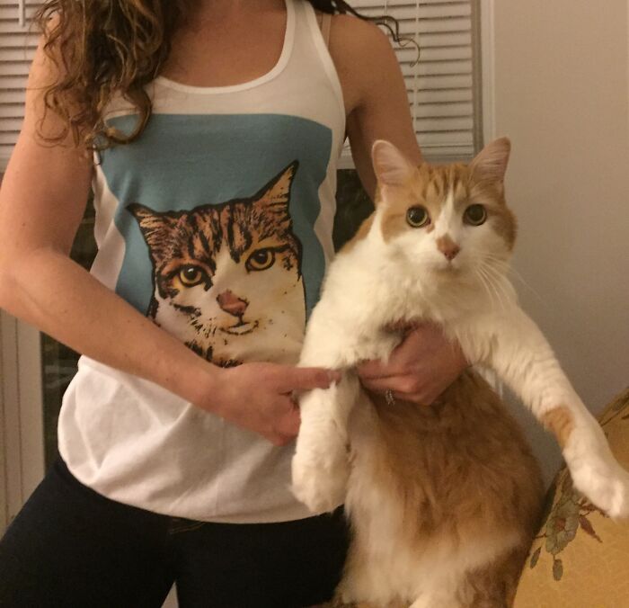 A woman in a tank top featuring a cat print holds an orange and white cat in her arms.